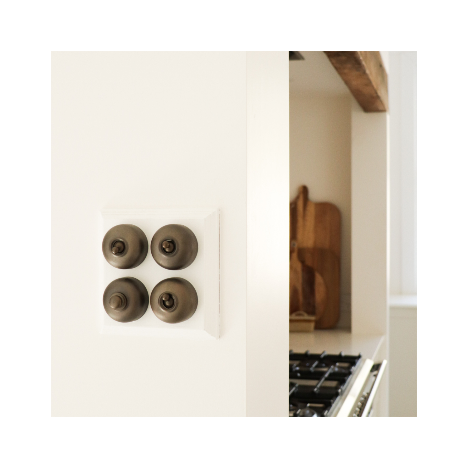 Traditional round antique brass toggle switches mounted on a white wall beside a kitchen opening, with a timeless white and timber kitchen visible beyond.