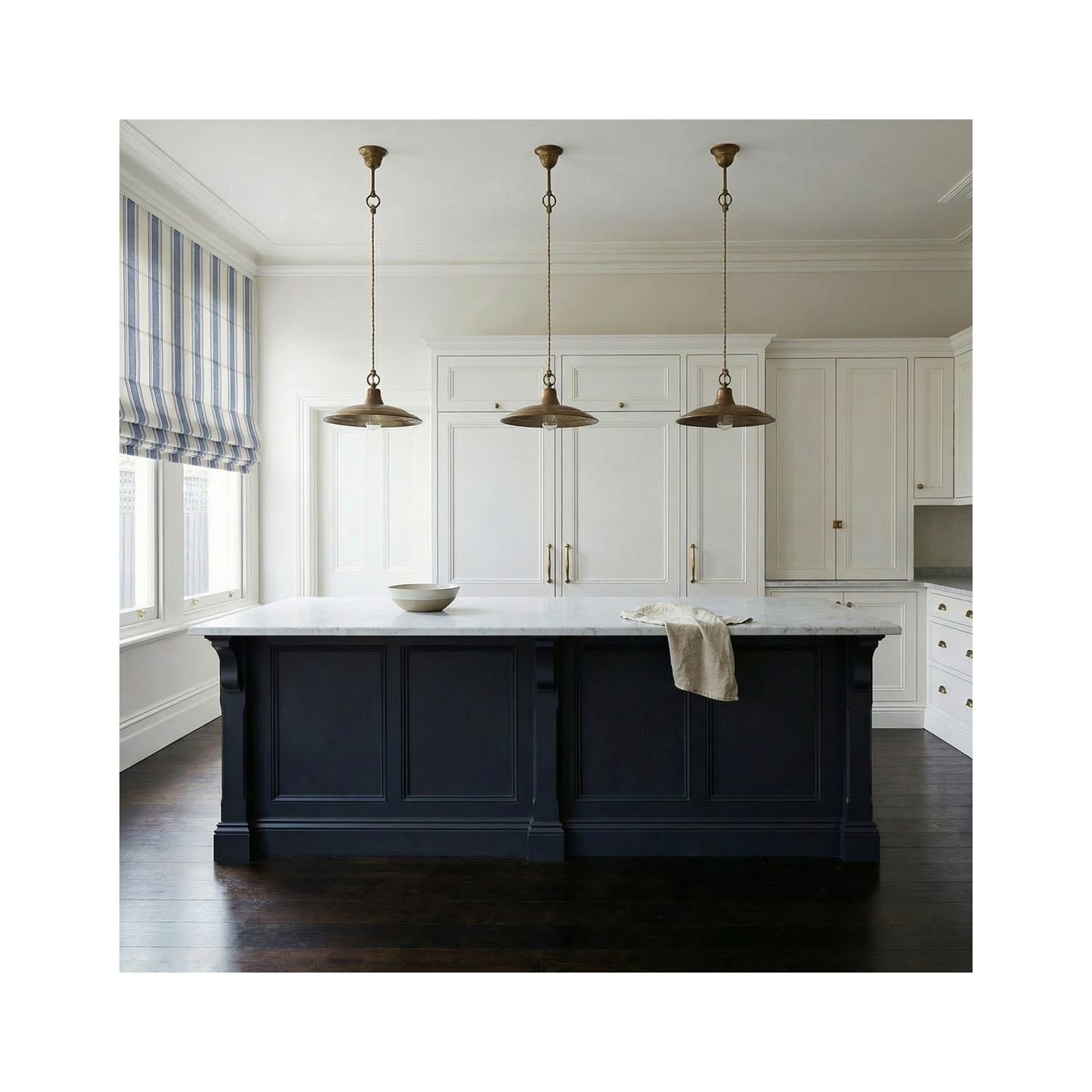 Three aged brass pendant lights hanging above a dark painted timber kitchen island with marble benchtop in a traditional period kitchen with sash windows and panelled cabinetry.