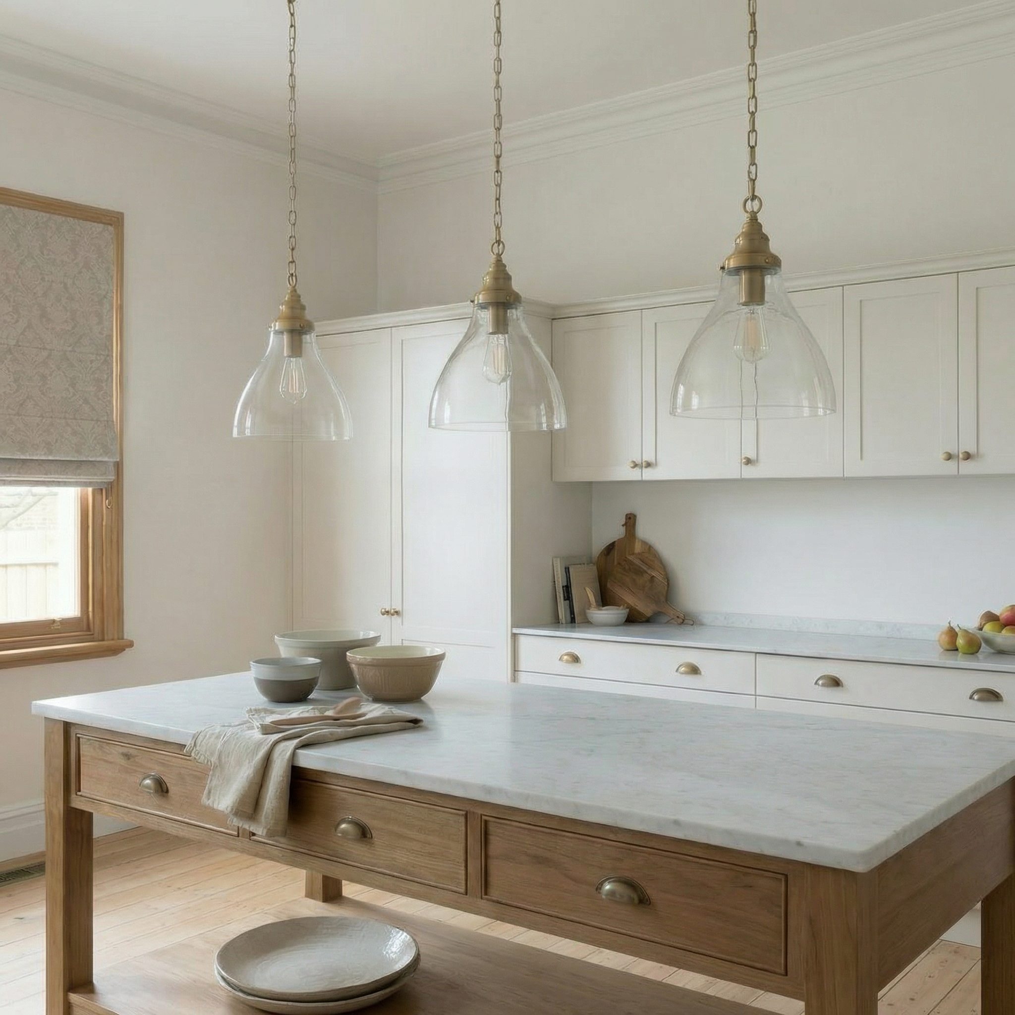 Three satin brass and clear glass pendant lights suspended over a timber island bench with marble top in a Federation-era kitchen featuring neutral white cabinetry, brass hardware, Tasmanian oak floorboards, and a Roman blind on a timber sash window.