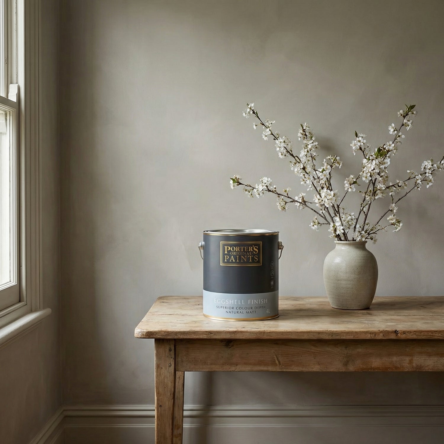 Porter’s Paints Eggshell Finish tin on a rustic timber table beside a ceramic vase of white blossom, set against a softly textured neutral wall in a period-style interior with natural daylight.